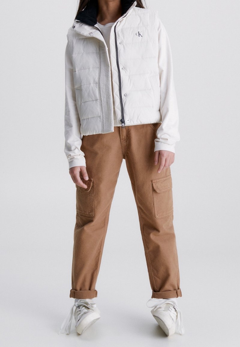 Weißer Puffer-Weste mit durchgehendem Reissverschluss, über einem weißen Langarmshirt getragen. Braune Cargo-Hosen mit aufgesetzten Taschen und weißen Turnschuhen.