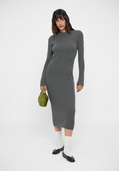 Robe en tricot côtelé gris avec des manches longues, une silhouette ajustée et un ourlet droit. Portée avec des chaussettes blanches et des ballerines noires, tenant un sac à main vert.