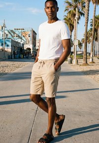 Bărbat îmbrăcat într-un tricou alb și pantaloni scurți bej stă pe o alee însorită, mărginită de palmieri, aproape de o plajă, cu clădiri urbane în fundal.