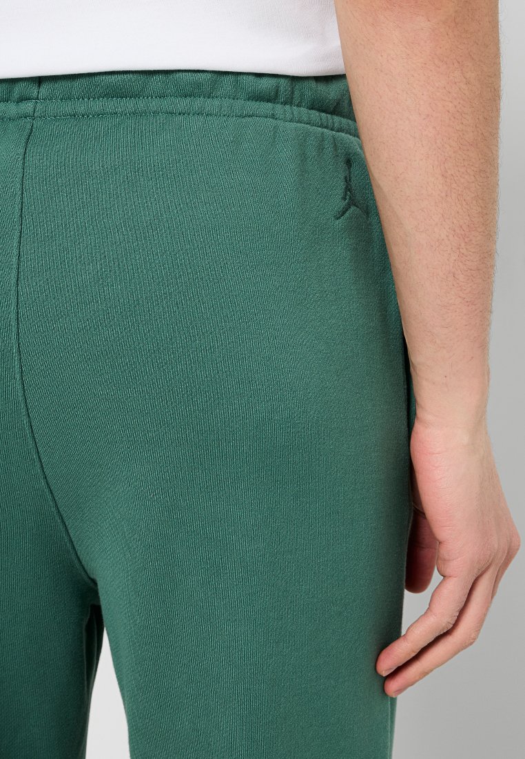 Pantalons de jogging en polaire verte avec une surface texturée, taille élastique, et un petit logo noir à l'arrière près de la taille, montrant un gros plan des détails.