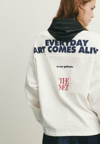 Jonge persoon met rood haar draagt een wit sweatshirt met een zwarte capuchon, met de tekst "EVERY DAY ART COMES ALIVE" en het logo van "THE MET" op de achterkant.