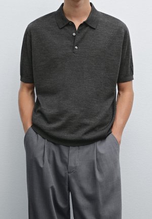 Homme portant un polo gris foncé à manches courtes et un pantalon gris plissé ample, les mains dans les poches, devant un mur uni.