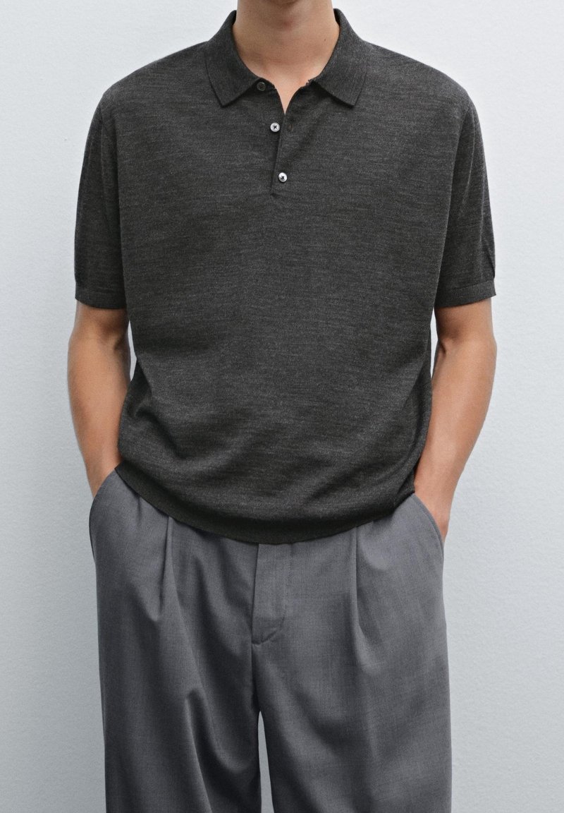 Homme portant un polo gris foncé à manches courtes et un pantalon gris plissé ample, les mains dans les poches, devant un mur uni.