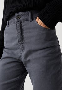 Szürke pamut nadrág sima textúrával, elülső zsebbel, gombos záródással és diszkrét varrási részletekkel. Fekete kötött pulóverrel viselve.