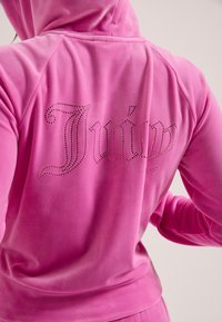 Femeie purtând un hanorac roz aprins cu litere gotice cu nituri pe spate și pantaloni roz asortați.