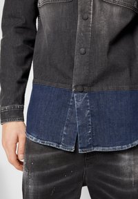 Giacca in denim nera con parte inferiore in denim blu. Presenta una chiusura a bottoni, texture a contrasto e un orlo leggermente curvo.