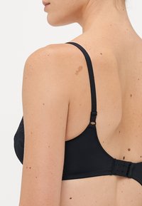 De rug van een vrouw met een zwarte beha met verstelbare bandjes en haak-en-oogsluiting, huid met sproeten en moedervlekken, op een effen lichte achtergrond.