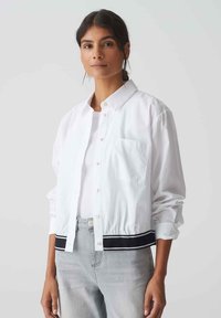 White button-up jacket with a collared neck, single chest pocket, and black striped hem. Light fabric with a relaxed fit and rolled sleeves.