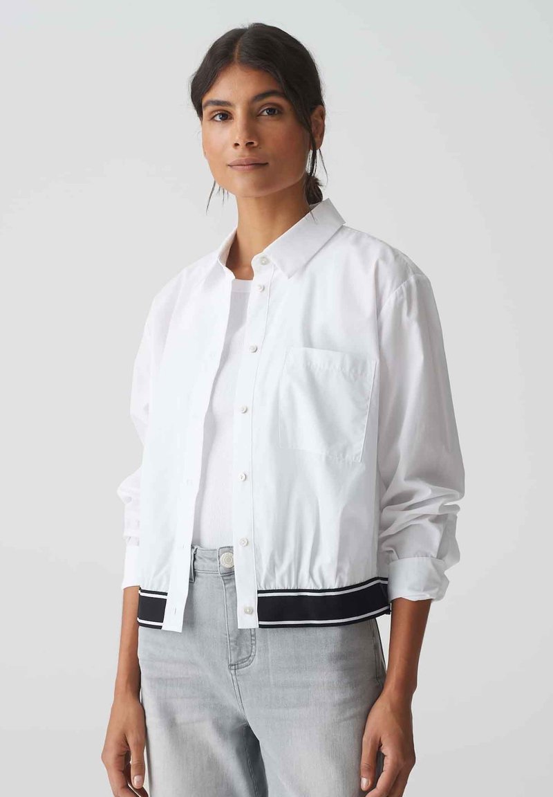White button-up jacket with a collared neck, single chest pocket, and black striped hem. Light fabric with a relaxed fit and rolled sleeves.