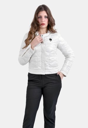 Giovane donna con lunghi capelli mossi indossa una giacca trapuntata bianca con bottoni a pressione e pantaloni neri, posando su uno sfondo semplice.