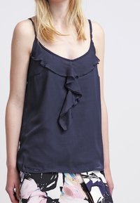 Granatowy top bez rękawów z falbanym dekoltem, wykonany z gładkiego, lekkiego materiału. Posiada cienkie ramiączka i luźny krój.
