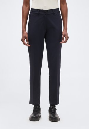 Persoon draagt slanke zwarte broek, zwarte veterschoenen en een lichtgekleurd bovenstuk, staand tegen een effen achtergrond.