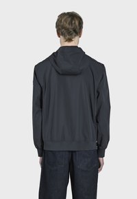 Chaqueta con capucha de color gris oscuro, fabricada con un material ligero. Cuenta con puños y dobladillo elásticos, y tiene un diseño minimalista. Se lleva con pantalones de mezclilla oscuros.