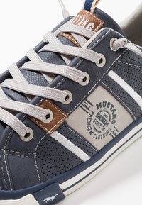 Granatowe trampki z szarymi akcentami, perforowaną teksturą i beżową naszywką z logo. Mają szare sznurowadła i biały wzór w paski.