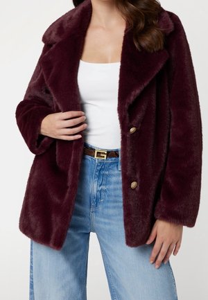 Femme portant un manteau en fausse fourrure bordeaux avec des boutons dorés par-dessus un top blanc et un jean bleu clair avec une ceinture à motifs.