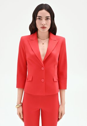 Donna con capelli lunghi e scuri che indossa un blazer rosso aderente con due bottoni, pantaloni rossi abbinati, braccialetti dorati e collane a strati.