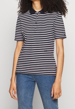Vrouw draagt een zwart-wit horizontaal gestreept kortärmelig poloshirt met zwarte kraag en blauwe spijkerbroek, staand tegen een neutrale achtergrond.