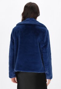 Kvinde if�ørt en mørkeblå blød faux fur-jakke set bagfra over en sort nederdel eller kjole.