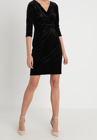 Svart sammetklänning med v-ringning, trekvartslånga ärmar och en samlad midjedetalj. Bärs med nude högklackade skor.