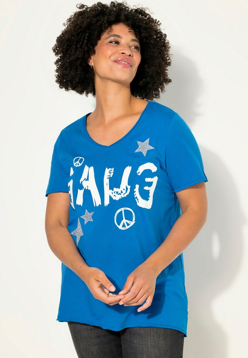 Camiseta de algodón azul con cuello en V, que presenta el texto impreso en blanco y plata "GANG" y signos de paz, así como patrones de estrellas. Mangas cortas.