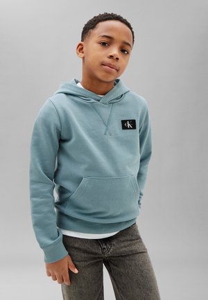 Chłopiec w jasnoniebieskiej bluzie z kapturem Calvin Klein i ciemnoszarych dżinsach stoi na jednolitym szarym tle, patrząc pewnie w kamerę.