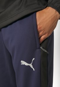 Námořnické atletické kalhoty s texturovanou tkaninou, s černým bočním panelem a stříbrným logem Puma. Obsahují detail s kapsou na zip.