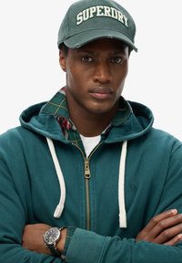 Sudadera con cremallera en color verde azulado y cordones blancos sobre una camisa de cuadros. Reloj negro en la muñeca. Gorra con el logotipo de "SUPERDRY" en blanco. Fondo uniforme.