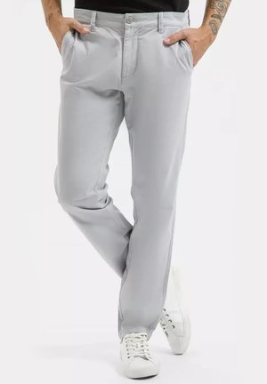 Jasnoszare casualowe spodnie noszone z białymi sneakersami, ręce w kieszeniach, jedna noga skrzyżowana na drugą, widoczne tatuaże na ramionach.