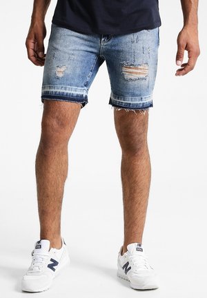 Vaaleansinisiä farkkushortsseja, joissa on kulutettu ilme, revitty helma ja klassinen viisi taskua -muotoilu. Yhdistetty valkoisiin lenkkareihin.