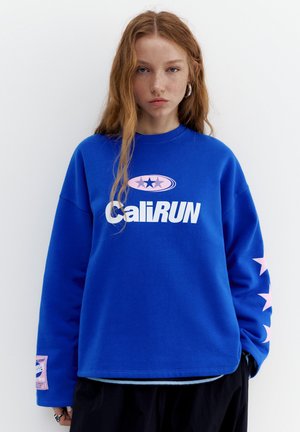 Mladá osoba s dlouhými rudými vlasy, která má na sobě modrou mikinu s nápisem "CaliRUN" a růžovými hvězdami na rukávu a na hrudi, stojící proti bílému pozadí.
