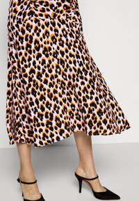 Jupe mi-longue à imprimé léopard orange, noir et blanc portée avec des escarpins noirs à bride arrière sur un fond clair.