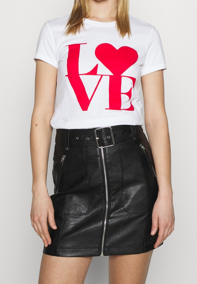 Femme portant un t-shirt blanc avec un texte rouge ""LOVE"" et un cœur, associé à une jupe en cuir noire taille haute avec fermeture éclair à l'avant et ceinture.