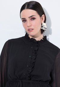 Černé šaty s volánkovým výstřihem a zapínáním na knoflíky. Dlouhé průsvitné rukávy, páskový pas a hladká textura. Velké kovové náušnice.