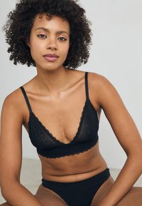 Black lace bralette with scalloped edges and thin straps, paired with matching black high-waisted underwear, set against a neutral backdrop.