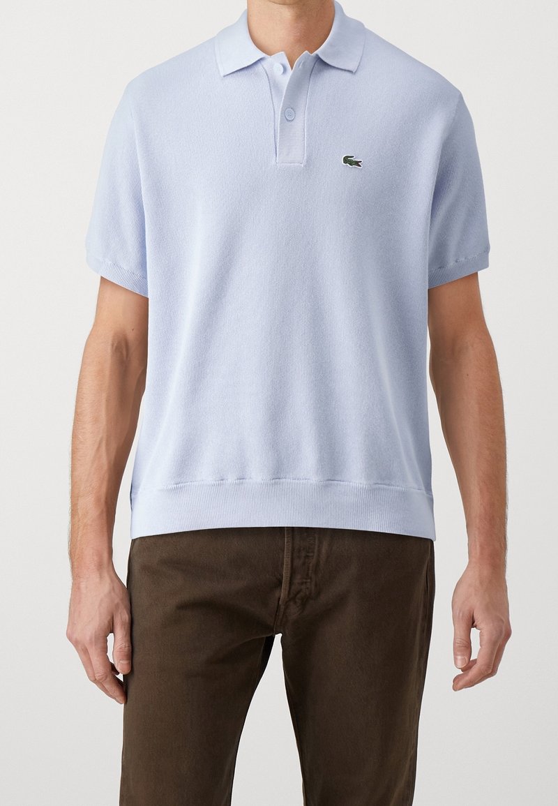 Ljusblå kortärmad polo med texturerad tyg; har en krage och broderad logotyp på bröstet.