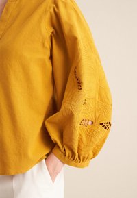 Blusa gialla senape con scollatura a V, maniche voluminose e ricami floreali intricati sui polsini. Realizzata in tessuto strutturato.