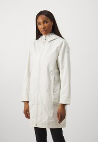 Lett, vanntett, lang hvit frakk med hette, frontglidelås, sidelommer og en litt blank tekstur. Enkel og funksjonell design.