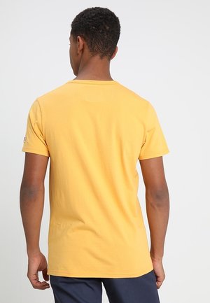 Mann trägt einfaches gelbes T-Shirt mit kurzen Ärmeln und dunkle Hose, steht mit dem Rücken zur Kamera vor einem schlichten hellen Hintergrund.