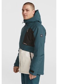 Chaqueta con cremallera en color verde azulado, negro y blanco, hecha de material impermeable, que incluye capucha, bolsillos con cremallera y puños ajustables.