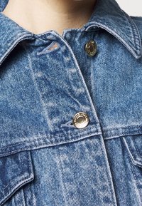 Veste en denim bleu clair avec un col classique, une fermeture à boutons dorés et des détails de couture visibles sur les poches et les coutures.