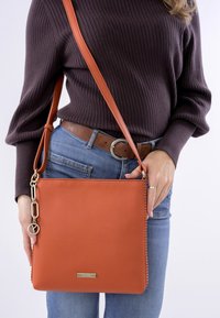 Orange leather crossbody bag with a smooth texture, rectangular shape, and metal hardware accent. Features a logo plaque and adjustable strap.