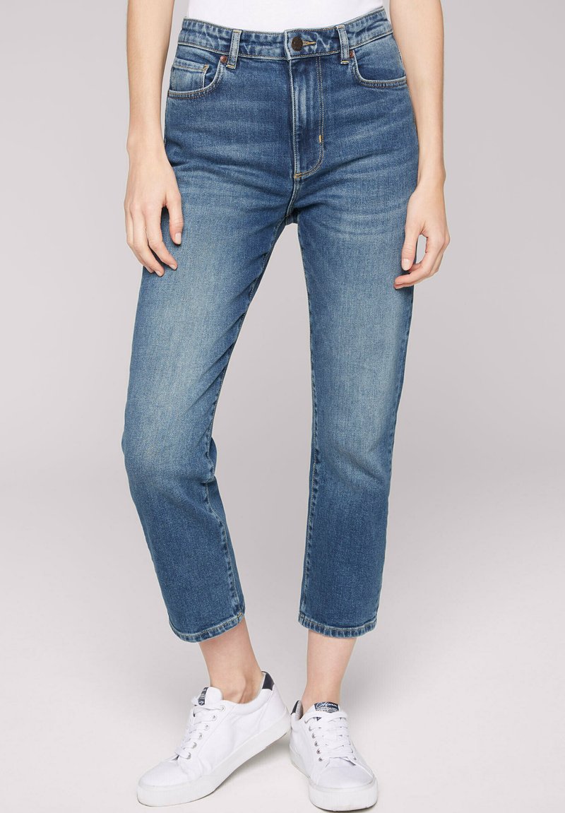 Personne portant un jean cropped bleu taille moyenne et des baskets blanches, se tenant devant un fond gris clair uni.