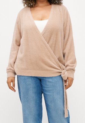 Kobieta w jasnobeżowym, puszystym swetrze zawiązanym z boku, nałożonym na biały top i niebieskie dżinsy, widoczna od pasa w górę na białym tle.