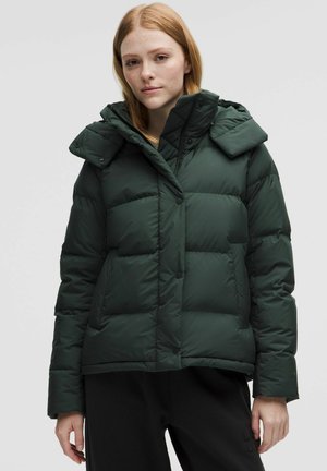 Giovane donna con capelli rossi che indossa una giacca imbottita verde scuro sopra pantaloni neri, in piedi davanti a uno sfondo luminoso e semplice.