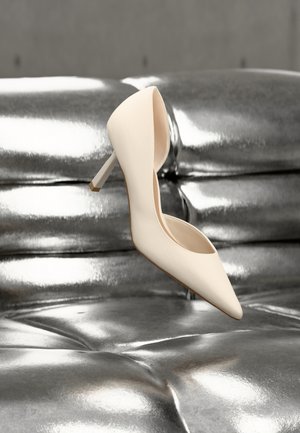 Cremefarbener High-Heel-Schuh schwebt über einem glänzenden silbernen, gesteppte Ledercouch, ohne sichtbare Personen oder Handlungen.