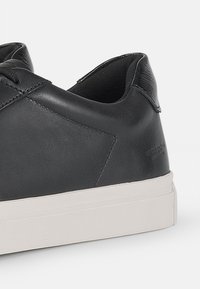 Sneaker in pelle nera con una texture liscia, suola in gomma bianca e design minimalista. Dettagli di cucitura evidenti e logo sul tallone.