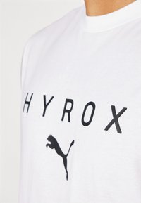 Biela bavlnená tričko s čiernymi tučnými písmenami hlásajúcimi "HYROX" a čiernym logom Puma. Dizajn s okrúhlym výstrihom, hladká textúra, štandardný strih.