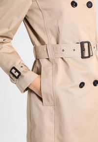 Beige trenčkot s dvojitými černými knoflíky, odpovídajícím páskem a nastavitelné manžety. Vyznačuje se hladkým, lehkým materiálem a bočními kapsami.