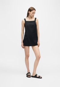 Jeune femme portant une robe courte noire sans manches et des sandales noires, debout devant un fond gris clair uni.