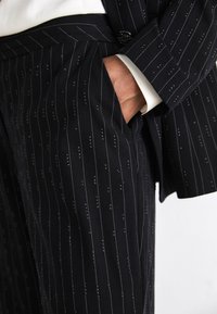 Costume noir sur mesure avec des rayures verticales blanches et de petites embellissements. Le tissu semble lisse avec un design structuré.